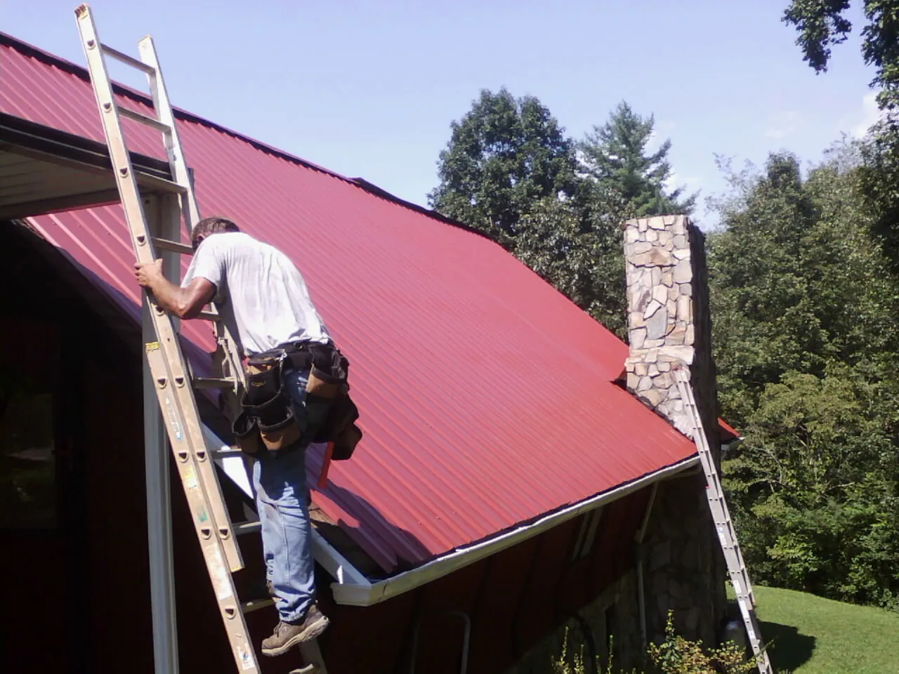 Roofer on a ladder beside a red metal roof and stone chimney — Soffit & Fascia Repair by Roofing Brothers in Parsonsburg